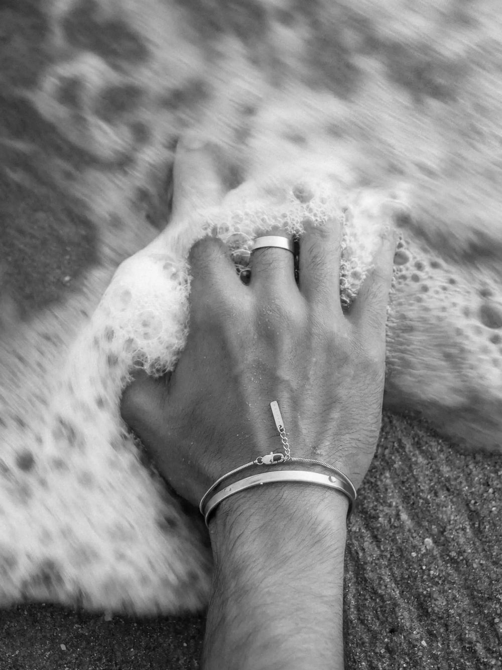Hand with a silver cuff, bracelet, and ring touching foamy water on a beach