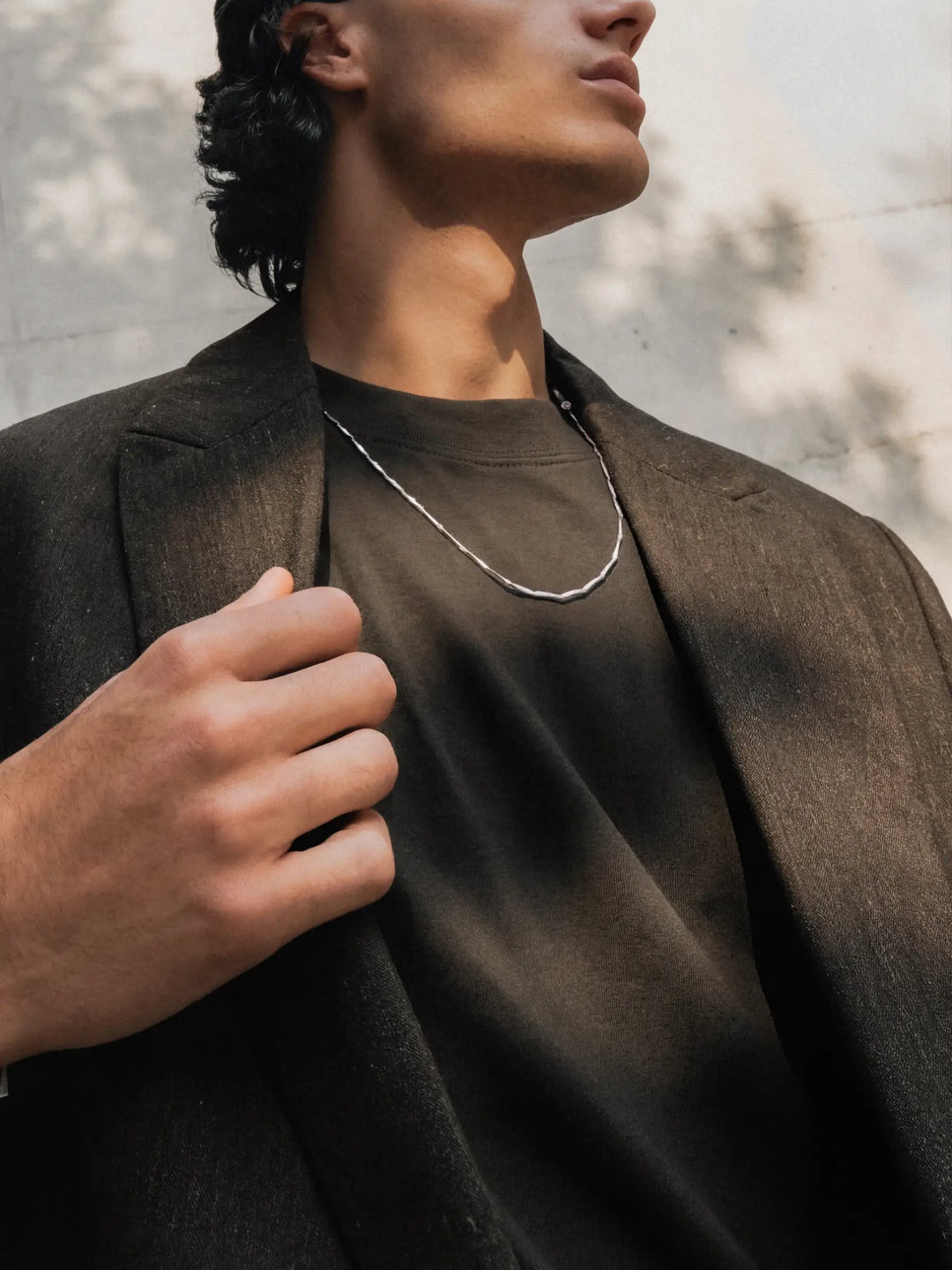 Person wearing a silver chain necklace with a dark coat and shirt with a neutral background