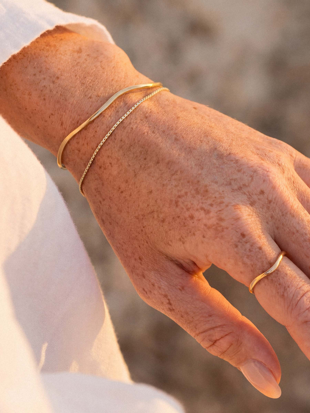 Close-up of a hand wearing a gold cuff, bracelet and ring against a blurred natural background