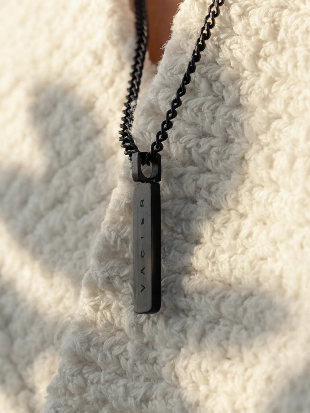 A close-up of a person wearing a black chain necklace with a rectangular pendant and a light-colored shirt