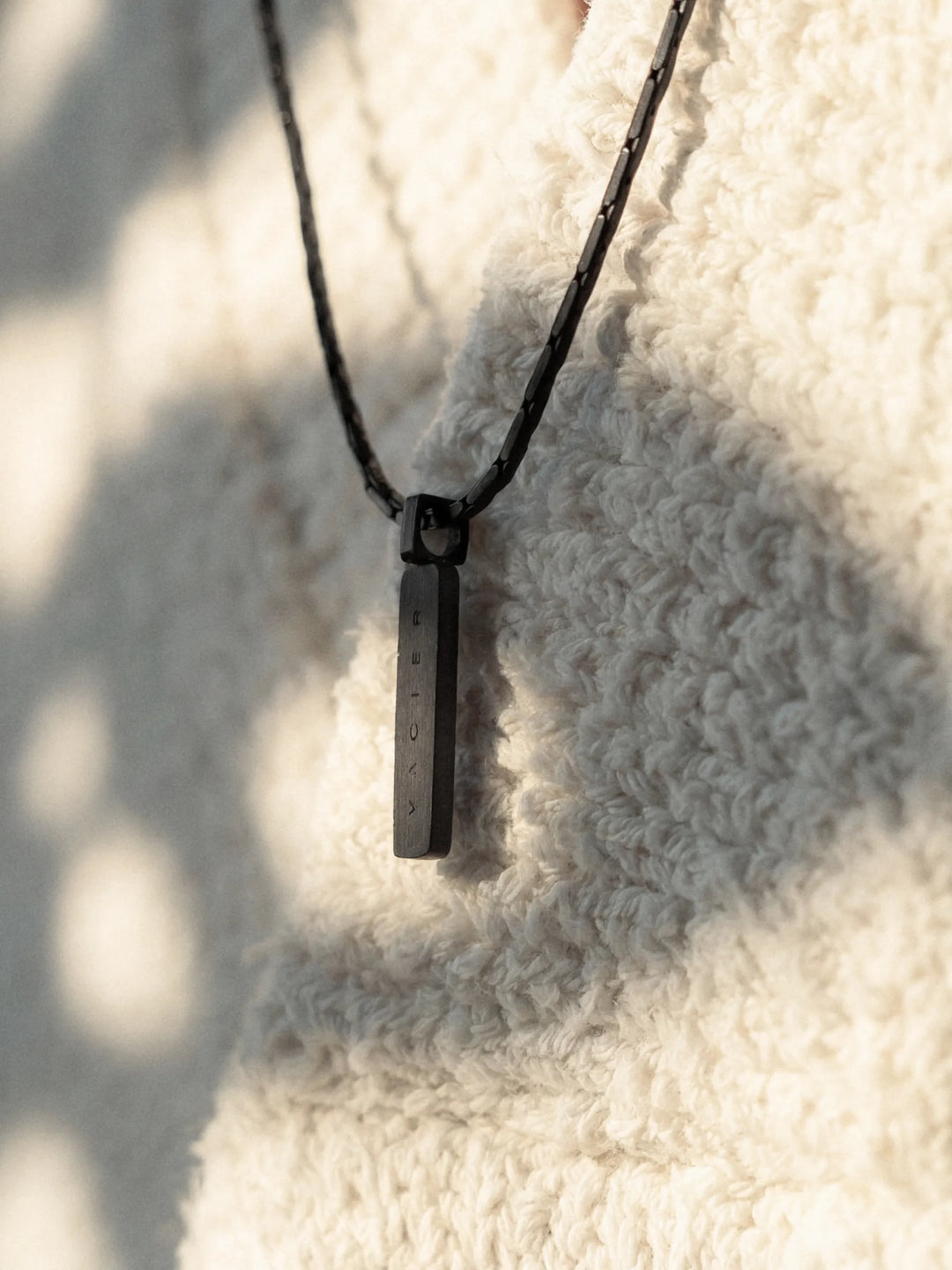 A close-up of a person wearing a black chain necklace with a rectangular pendant and a light-colored shirt