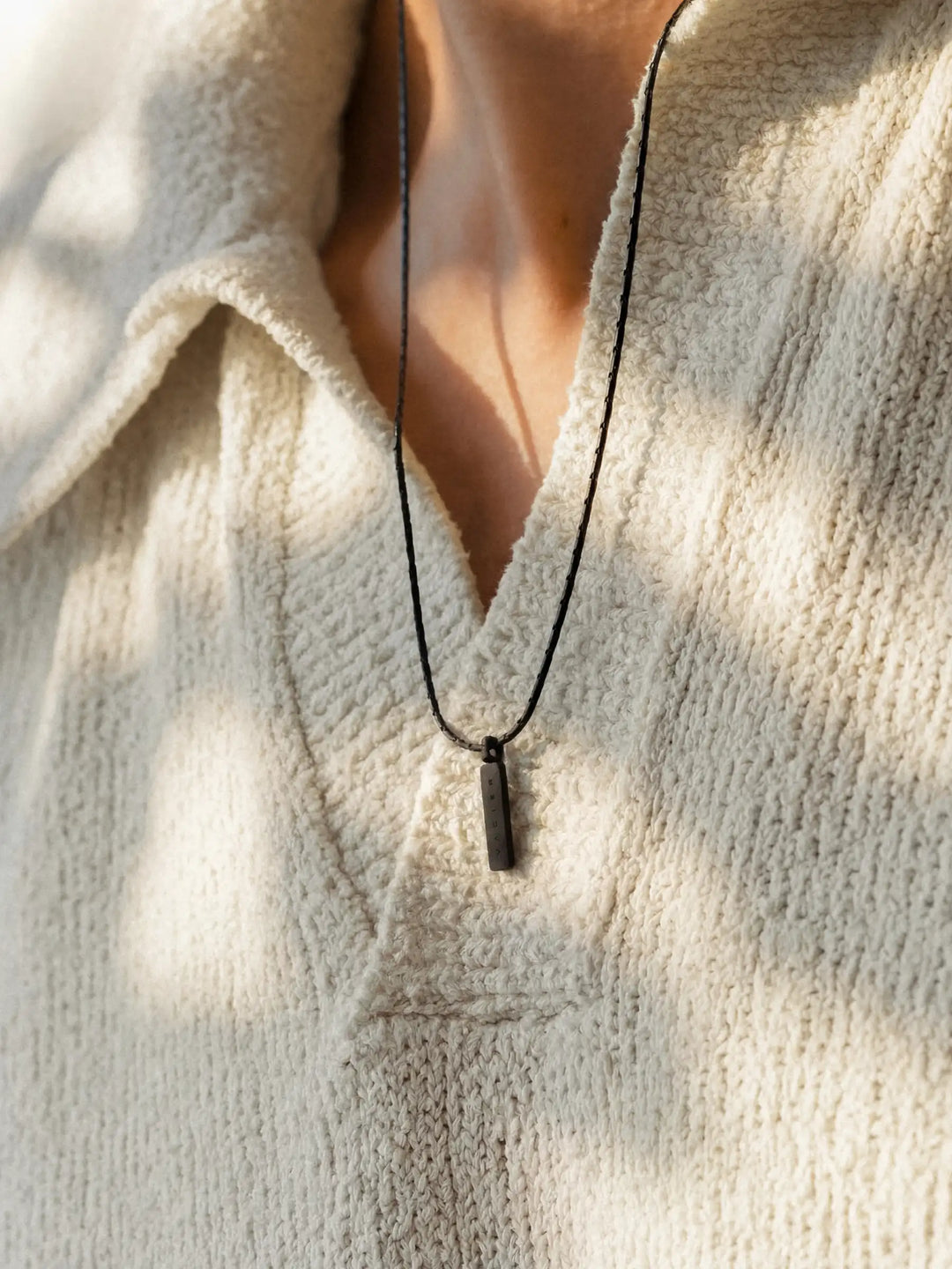 A close-up of a person wearing a black chain necklace with a rectangular pendant and a light-colored shirt