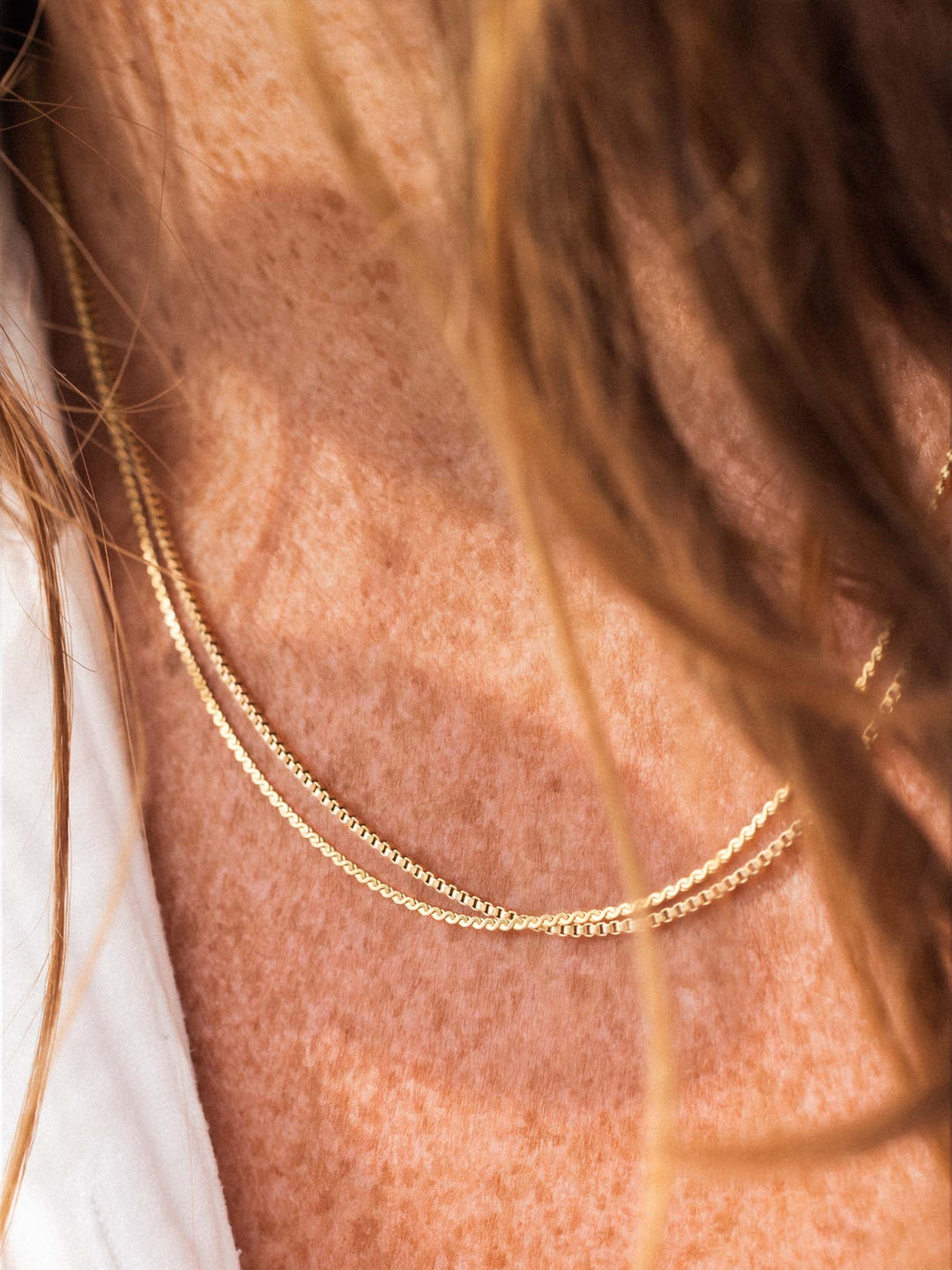 Close-up of a gold and a silver necklace on a person's neck with blurred hair in the foreground