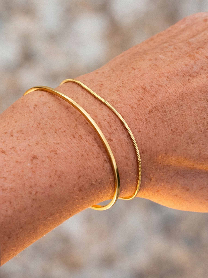 Gold bangle and a gold bracelet on a wrist with a blurred natural background