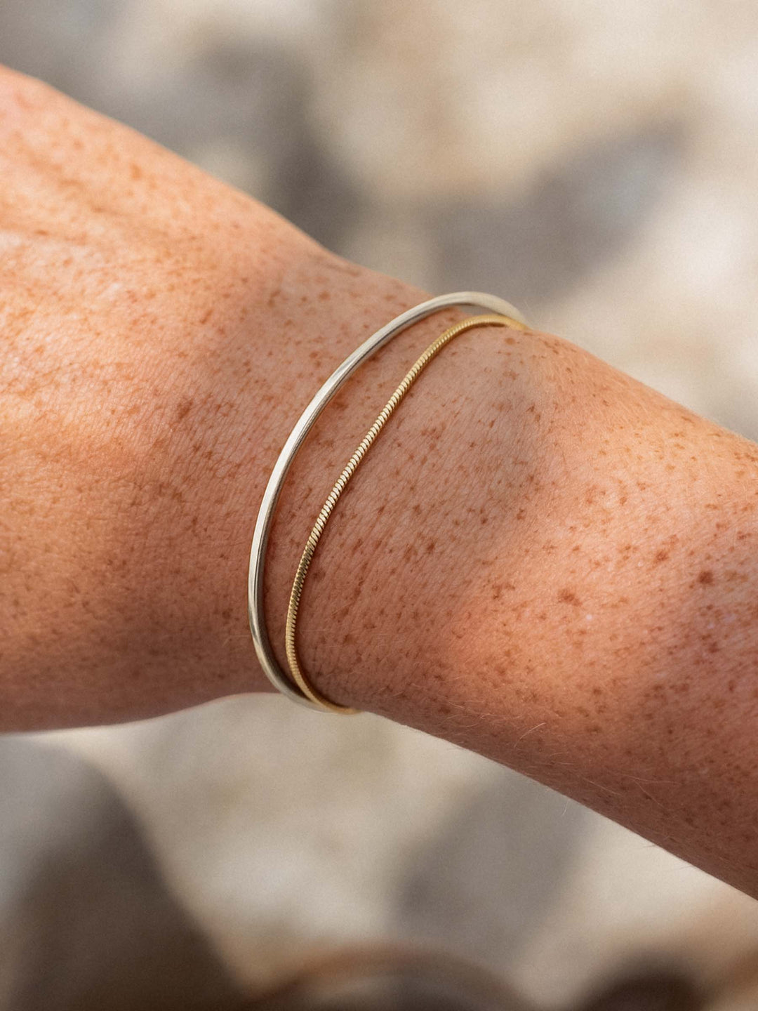 Close-up of a person's arm wearing a gold and a silver bracelet with a blurred natural background.